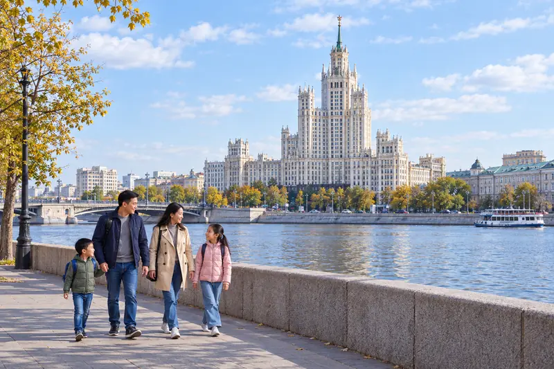 Gia đình Việt bốn người đi dạo trên bờ kè đá granite sông Moscow, phía bên kia sông là toà nhà chọc trời kiểu Stalin màu kem có chóp nhọn