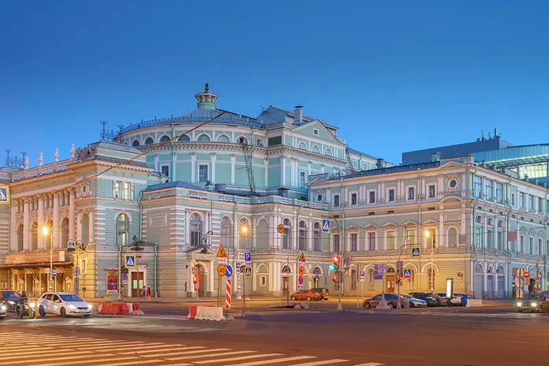 Nhà hát Mariinsky cũ ở St. Petersburg: toà nhà sơn xanh ngọc nhạt với mái vòm và cột trắng trên quảng trường Nhà hát, bên phải phía sau là khối kính của Mariinsky-2, đường phố và ô tô phía trước