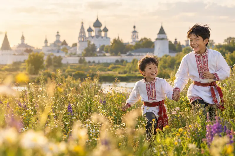 Hai anh em trai người Việt mặc áo thêu trắng thắt lưng đỏ nắm tay chạy giữa đồng hoa lúc hoàng hôn, xa xa là tường trắng và mái vòm tu viện Suzdal