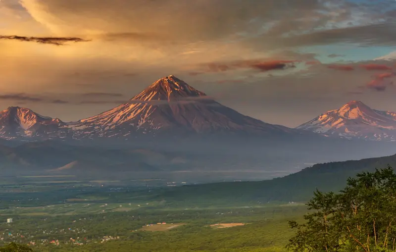 Núi lửa hình nón phủ tuyết của Kamchatka dưới bầu trời chiều, thung lũng xanh phía trước
