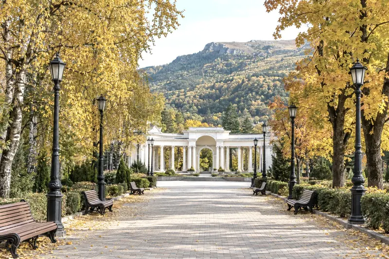 Đại lộ đi bộ trong công viên nghỉ dưỡng Kislovodsk, hàng cây vàng, cột đèn gang và ghế gỗ, hành lang cột trắng cuối đường, đồi núi phía xa